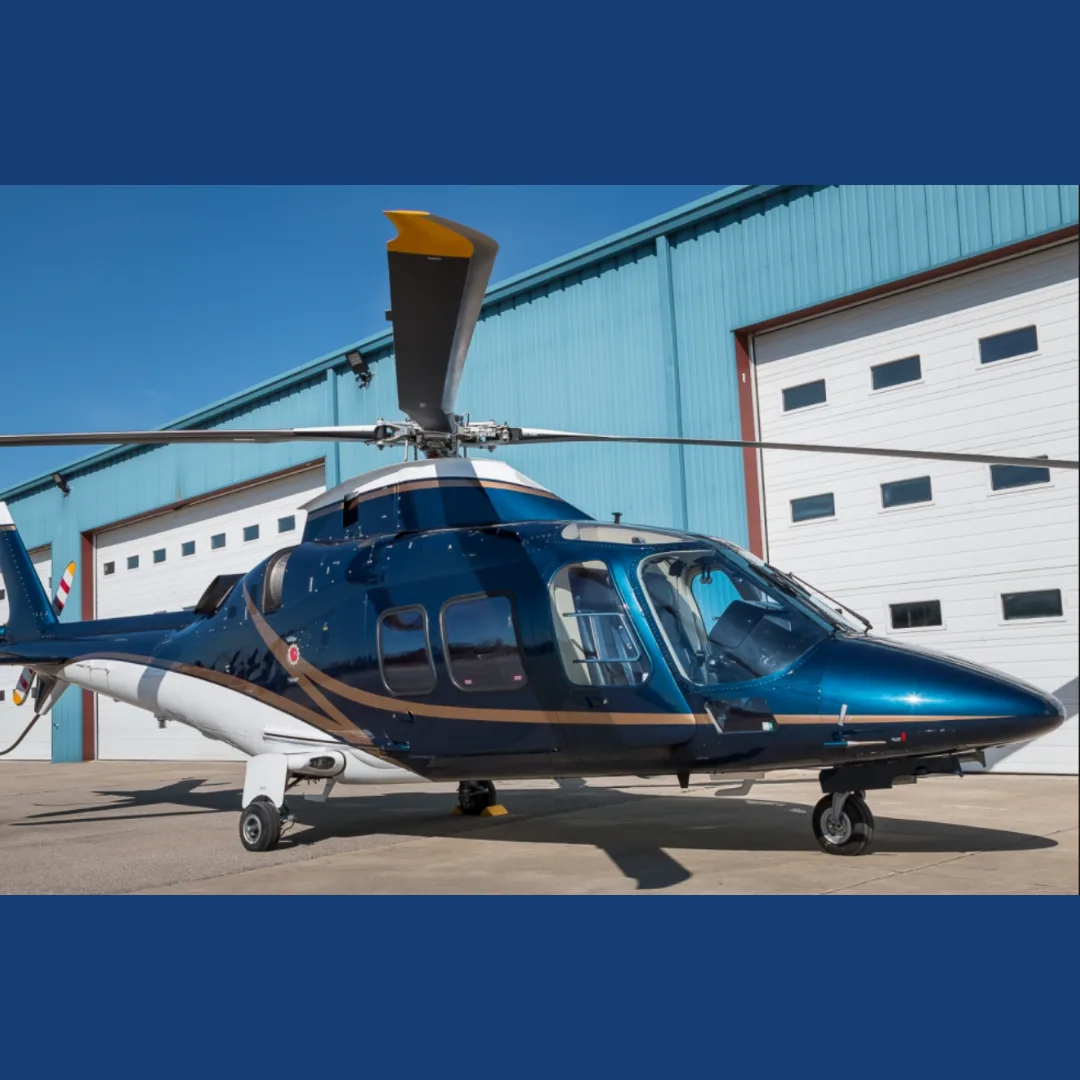 A dark blue and white helicopter with gold accents is parked on a tarmac in front of a light blue hangar under a clear sky.