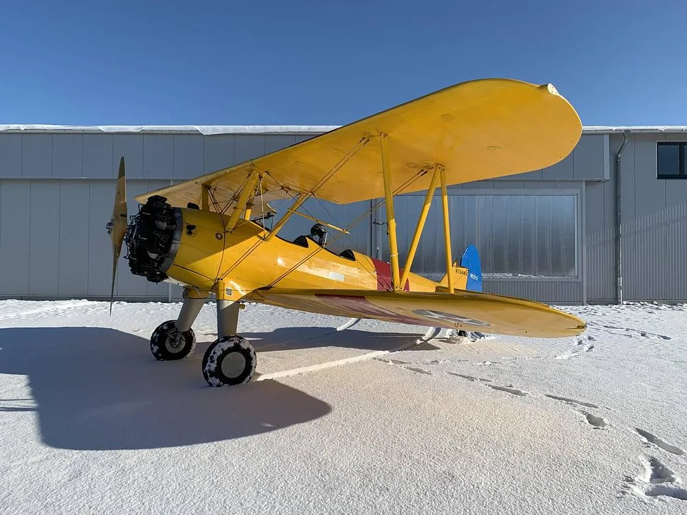 A yellow Stearman biplane with red and blue accents, marked N1344V, parked in the snow next to a hangar.