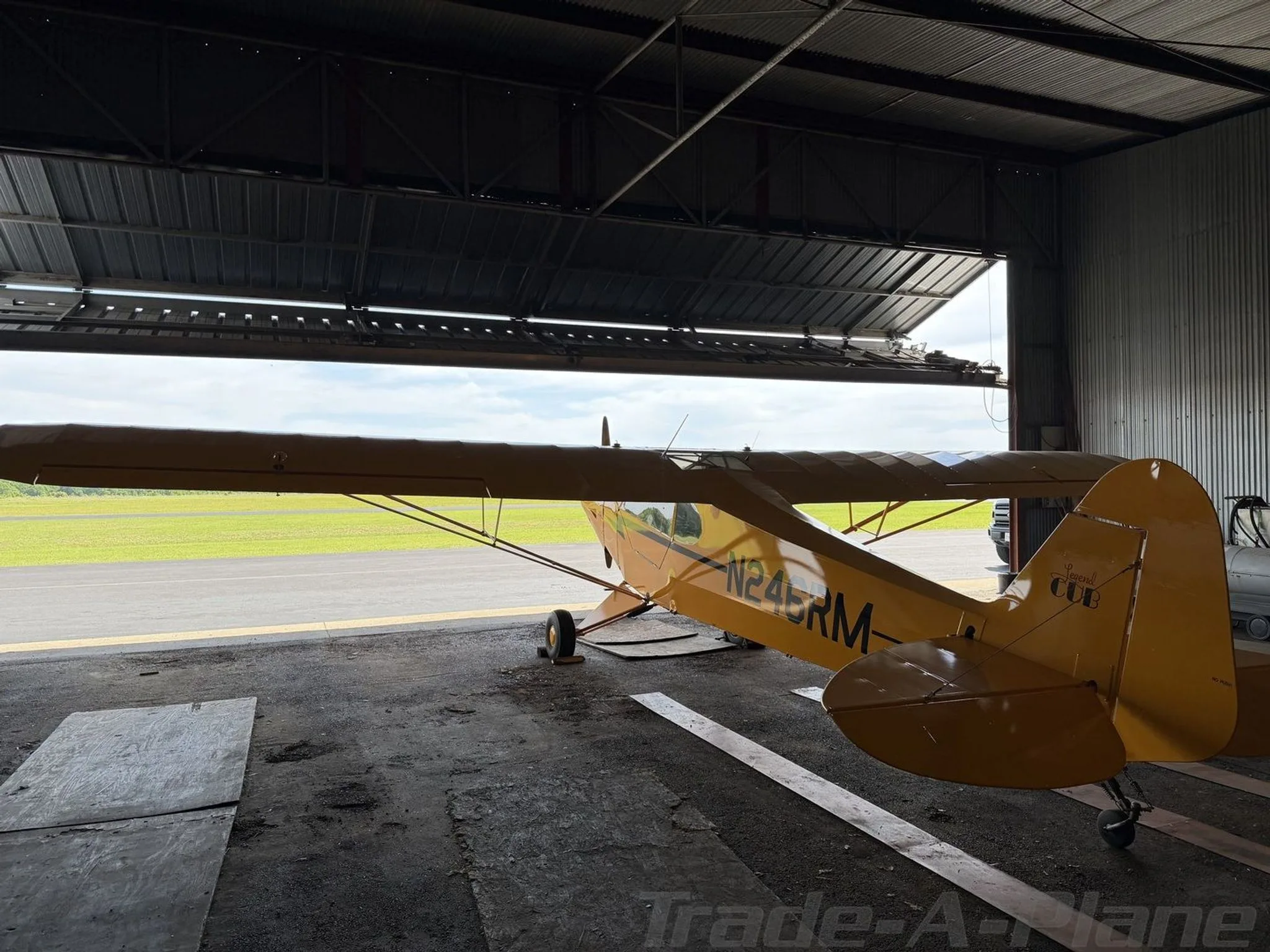 Rear-side view of a yellow American Legend Cub aircraft in a hangar, looking out to a green airfield.