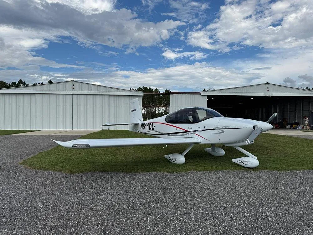 A white Van's RV-10 aircraft with red and black accents, registration N511DL, parked on grass next to hangars under a partly cloudy sky.