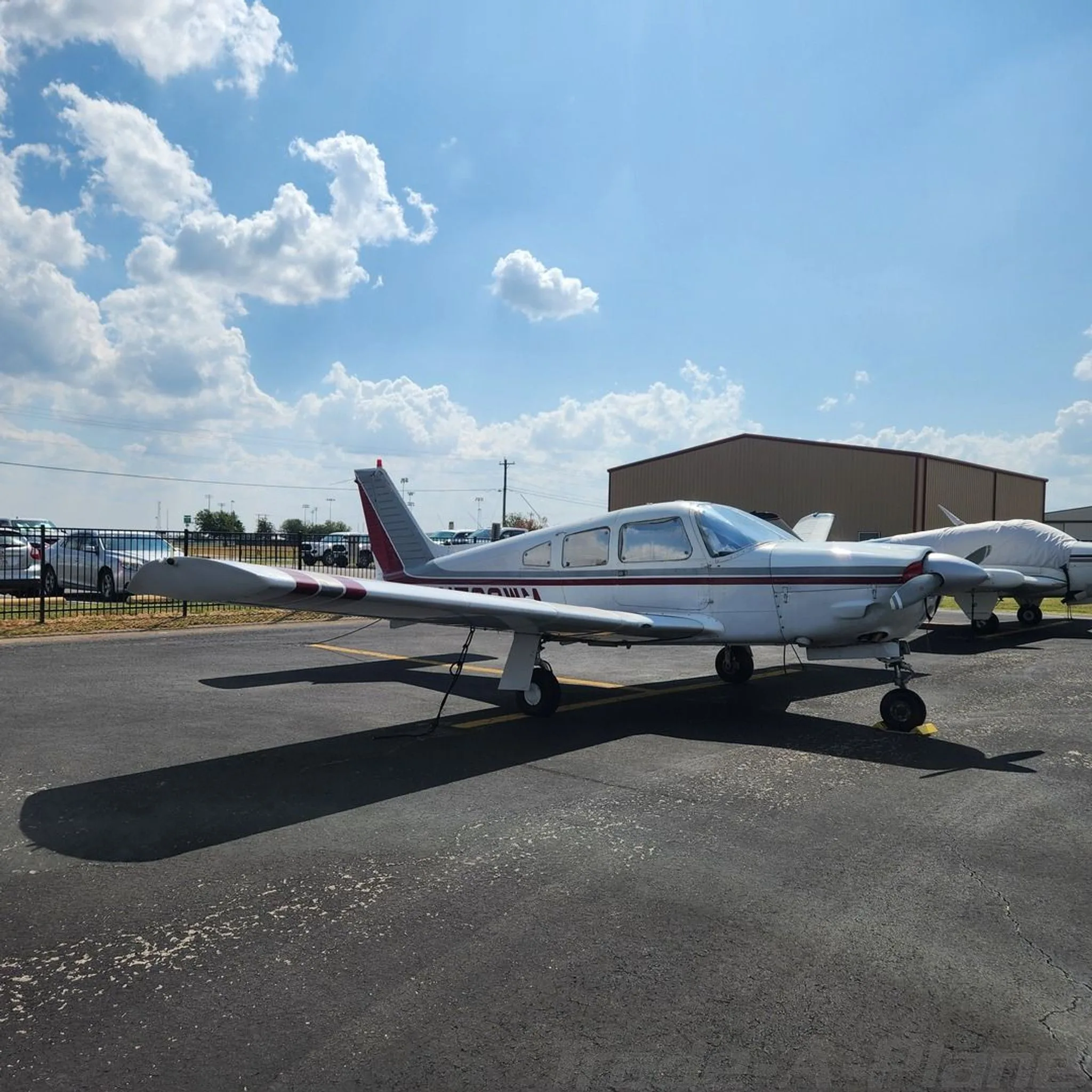 A white Piper Cherokee with red and grey stripes, registration N722WM, parked on an asphalt ramp with a fence and cars in the background.
