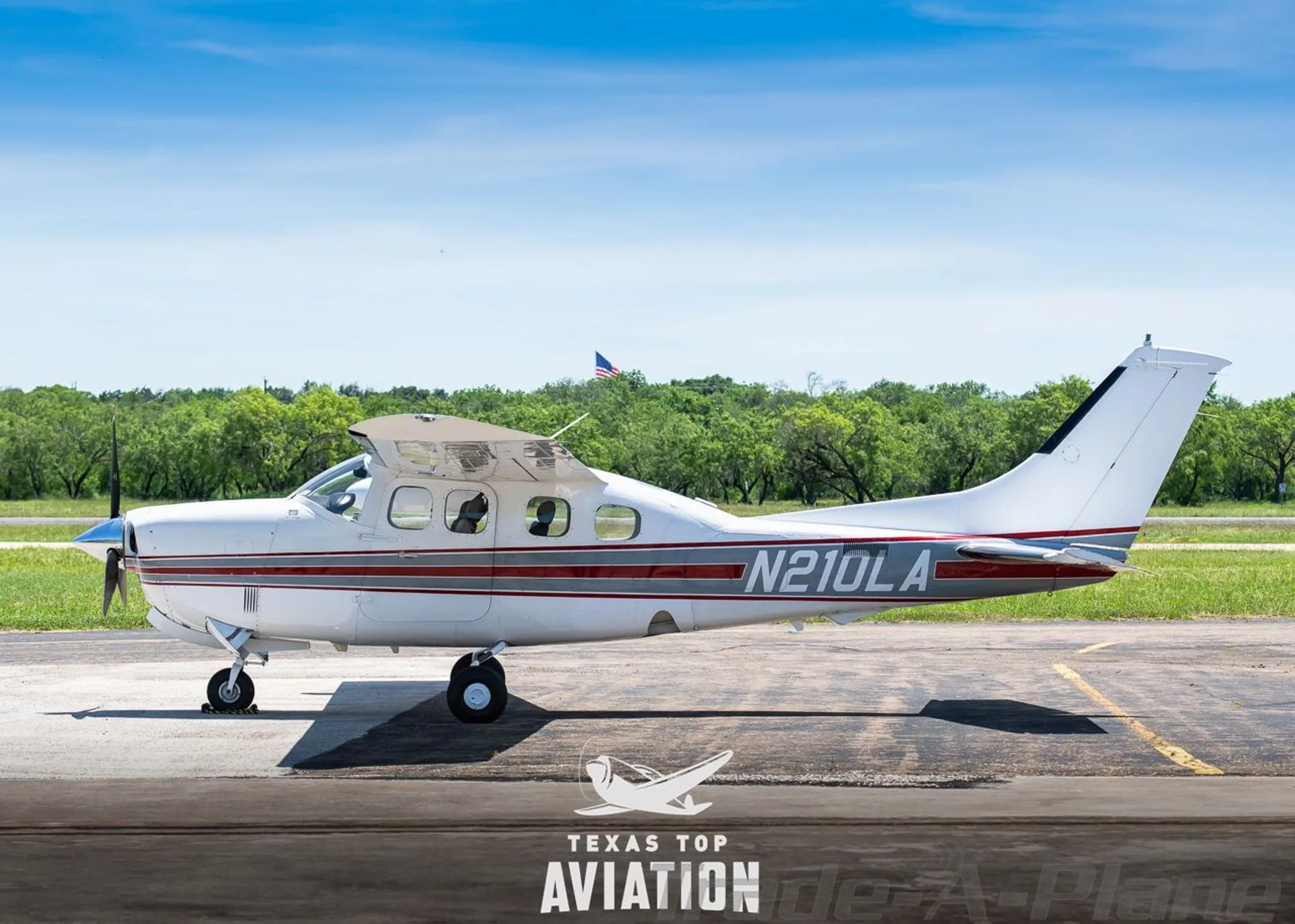 A white Cessna 210 Centurion (N210LA) with red and gray stripes parked on an airfield, showing the full side profile.