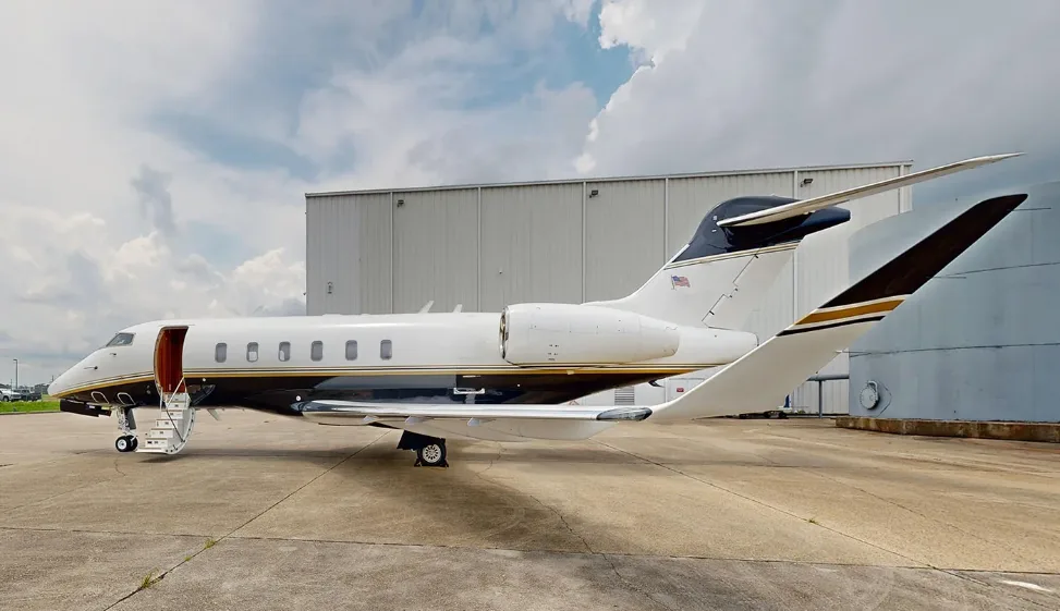 A white and black private jet with an open door and stairs on a tarmac next to a hangar under a cloudy sky.