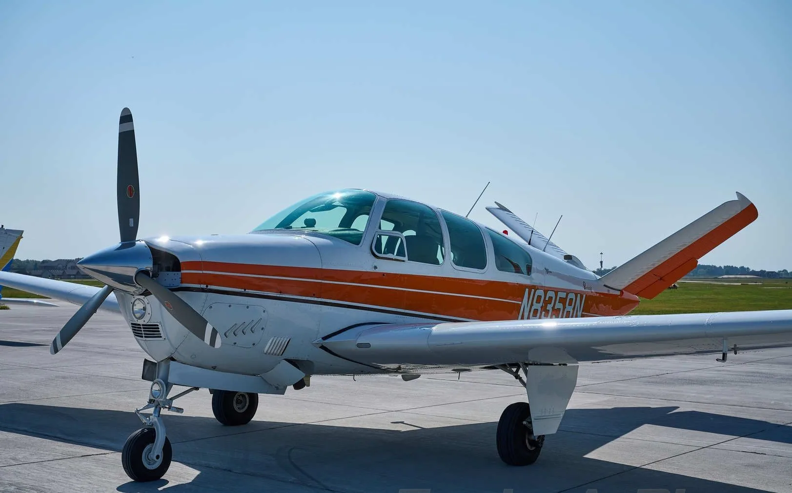 A white and orange Beechcraft Bonanza with a V-tail is parked on a tarmac under a clear blue sky.