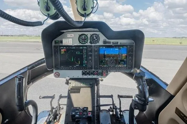 The cockpit of a helicopter showing a dual-screen glass panel display and flight controls.