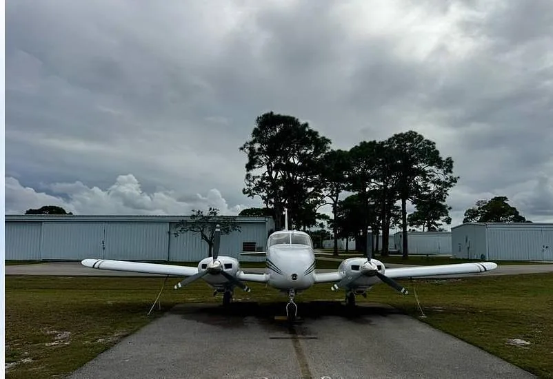 A head-on view of a white twin-engine aircraft with blue and gold stripes, parked on an asphalt strip at an airfield under a cloudy sky.
