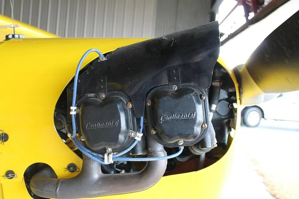 A close-up view of the Continental engine on the yellow aircraft, showing the exhaust pipe and blue ignition wires from a slightly different angle.