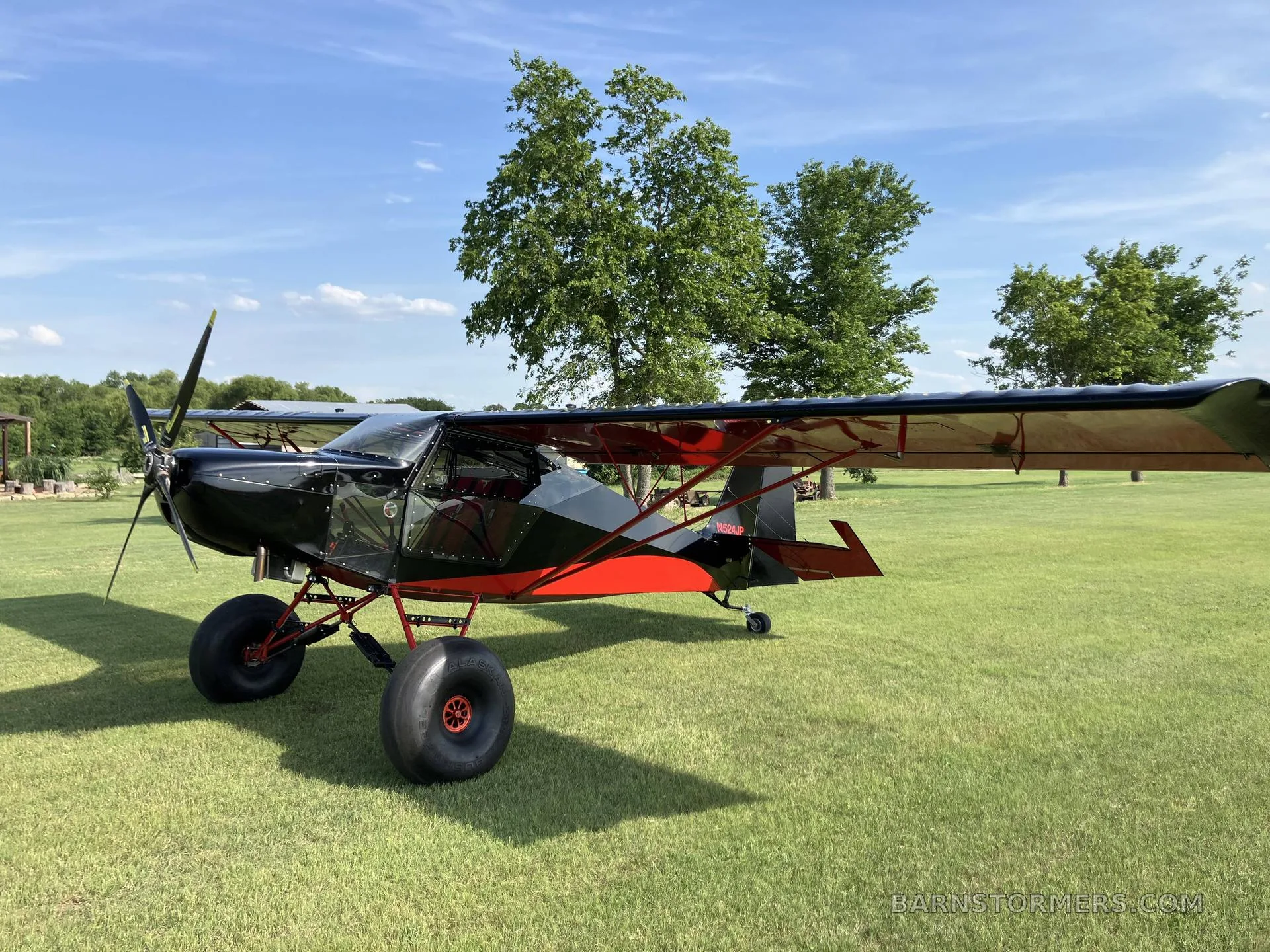 A black and red bush plane with large tires is parked on a grassy field under a blue sky.