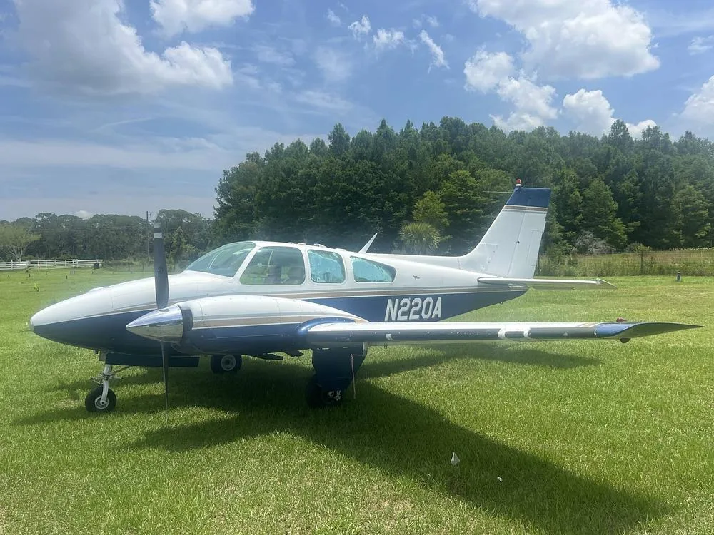 A white and blue twin-engine aircraft, N220A, is parked on a grassy field under a partly cloudy sky.