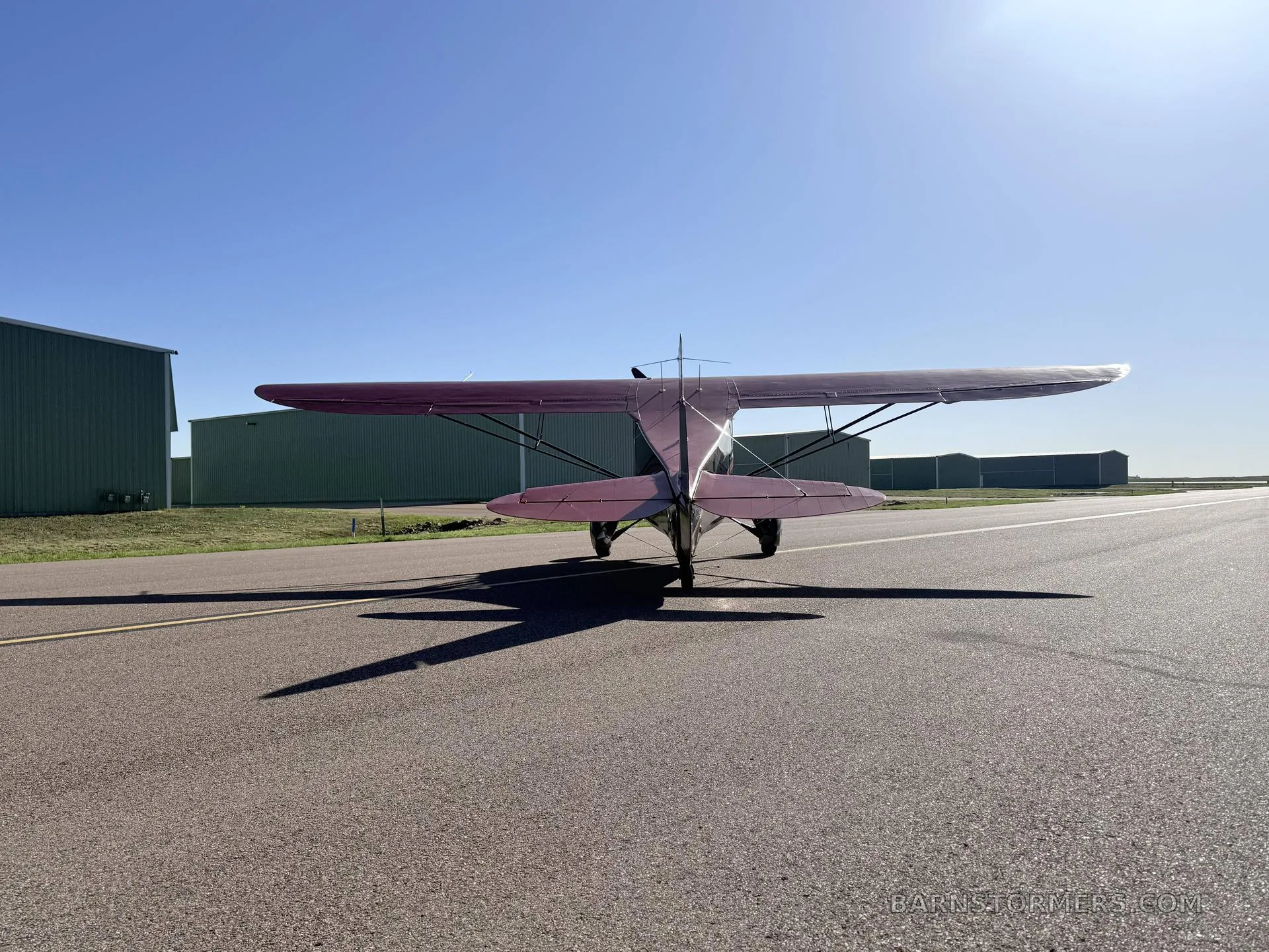 A rear view of a black and red vintage high-wing monoplane with tail number N999WT, casting a long shadow on an asphalt runway.