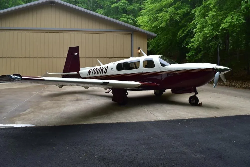 A maroon and white single-engine aircraft, N100KS, parked on a concrete pad in front of a light brown hangar.