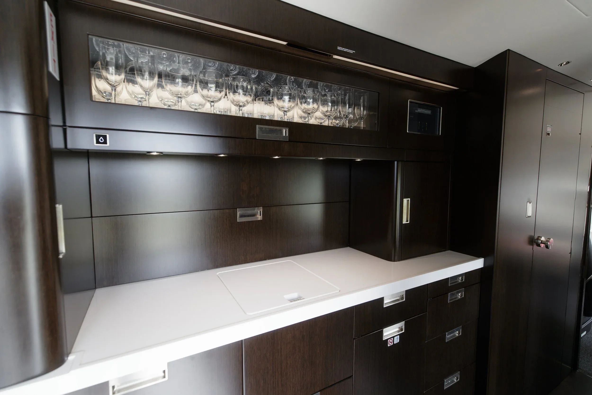 The dark wood galley of a private jet, featuring a white countertop, sink, and glass storage.