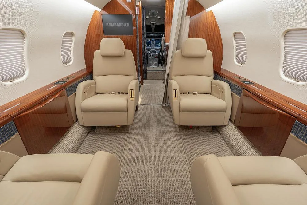 The interior cabin of a business jet, looking forward towards the galley and cockpit entrance, with beige seats and wood trim.