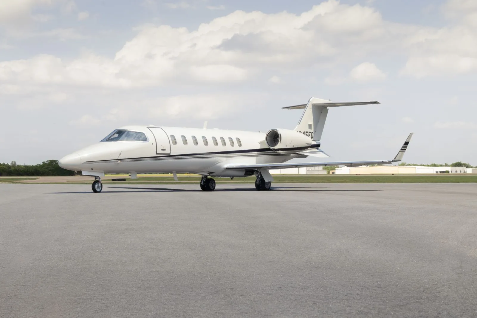 A white Learjet 45 business jet with black accent stripes is parked on an asphalt tarmac under a partly cloudy sky.