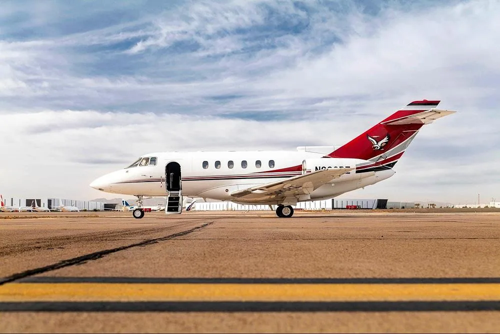 A white business jet with red and black stripes and an open door on a tarmac under a partly cloudy sky.