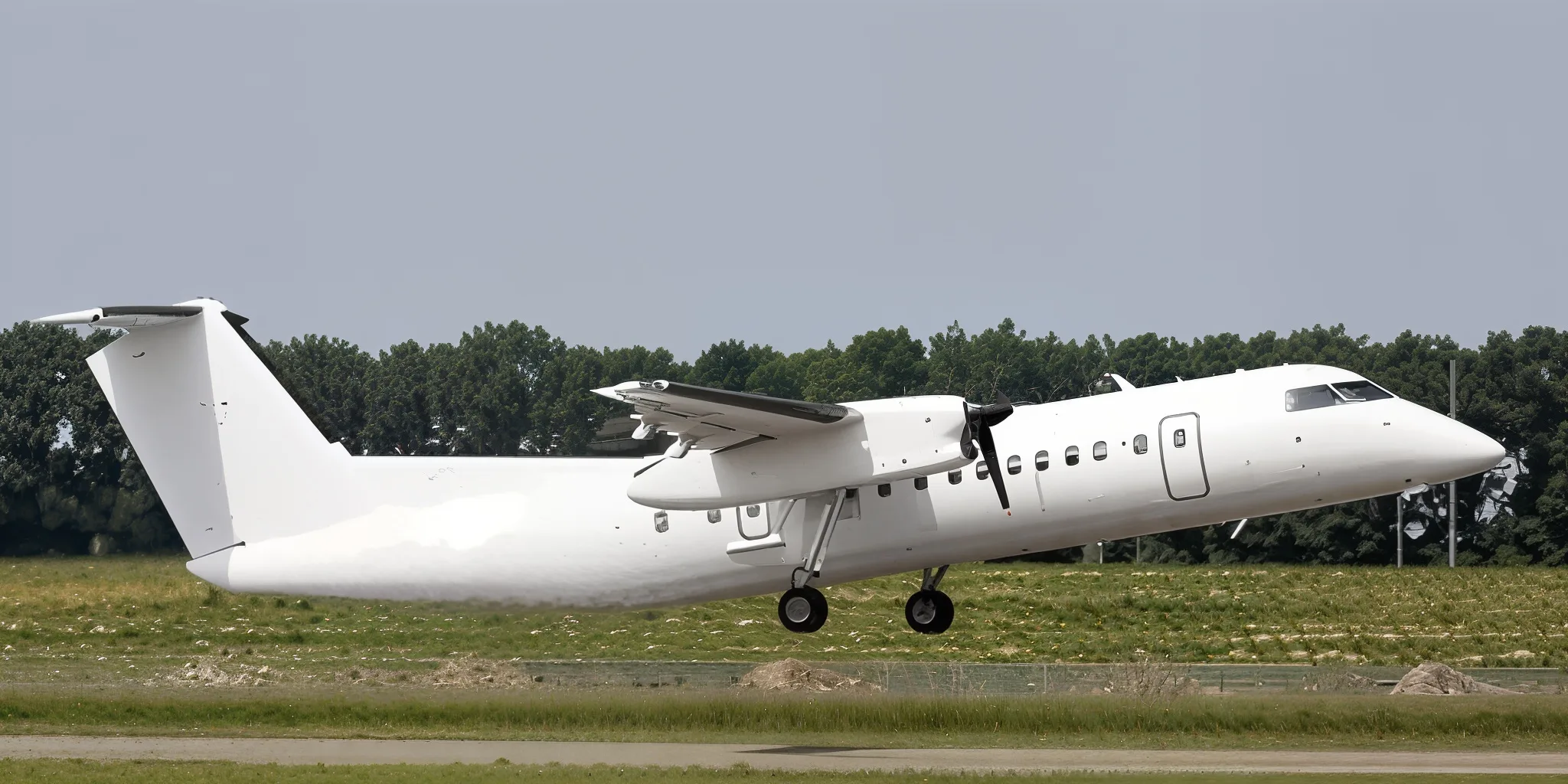 A white turboprop aircraft with landing gear extended, flying low over a grassy airfield.