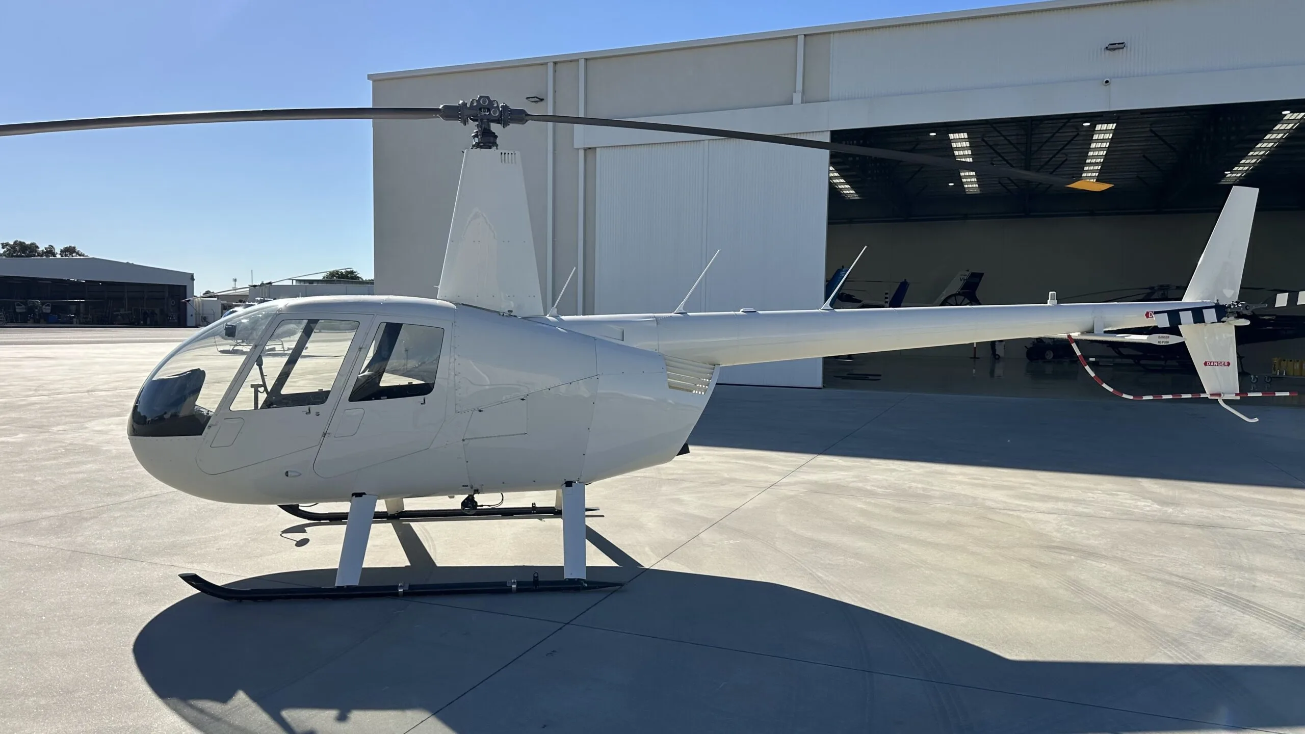 A white Robinson R44 helicopter parked on an airfield tarmac in front of an open hangar.