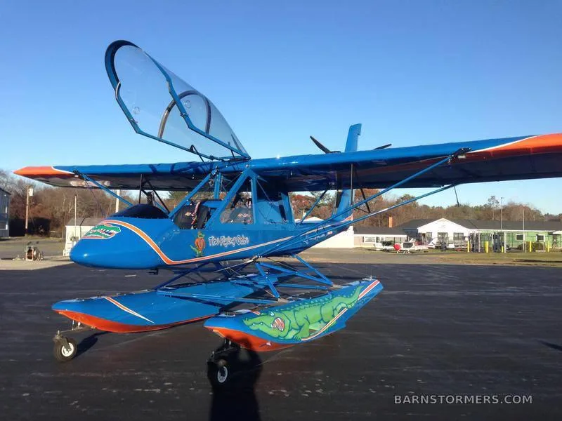 A blue floatplane with "The Flying Gator" and an alligator graphic on its floats, viewed from a slightly different angle on an tarmac.