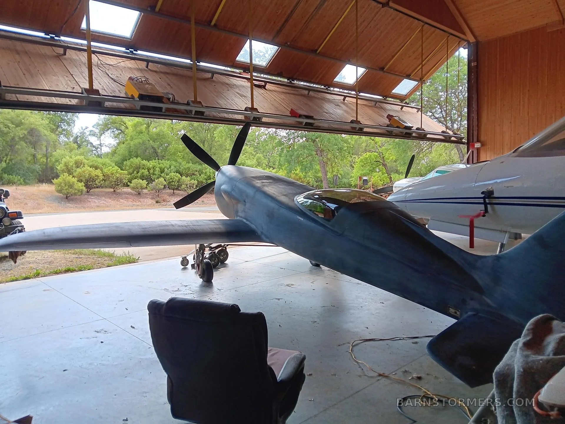 A dark, single-engine aircraft with a propeller, viewed from the side-rear, parked inside a hangar with an open door.