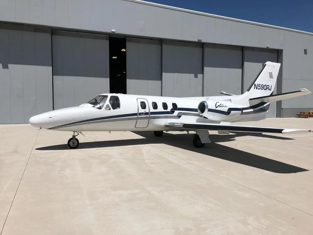 A white Cessna Citation jet with dark blue and grey stripes, registration N590RJ, parked on an apron in front of a hangar.