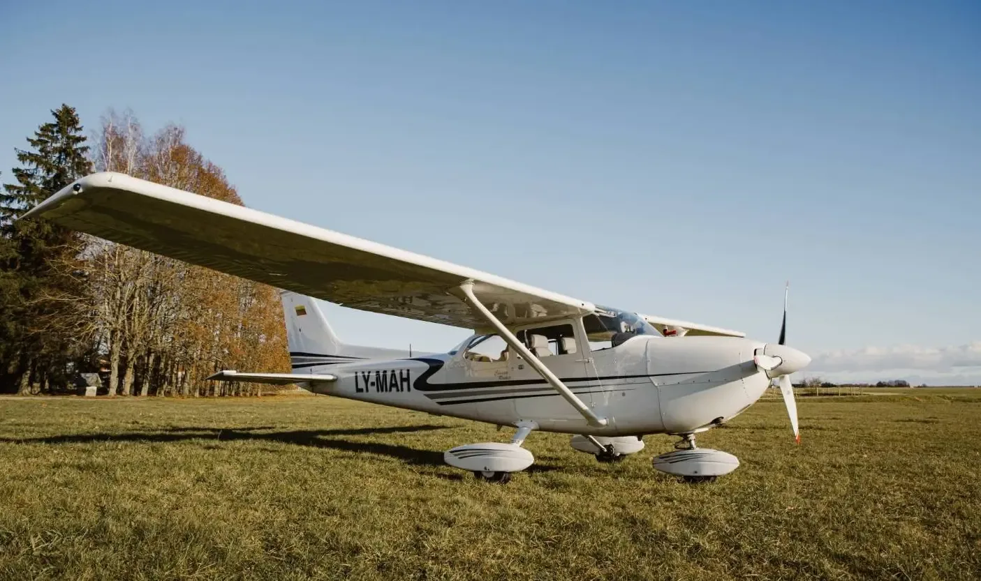 A white Cessna 172 with registration LY-MAH, viewed from the front-right, parked on a grassy field with trees in the background.