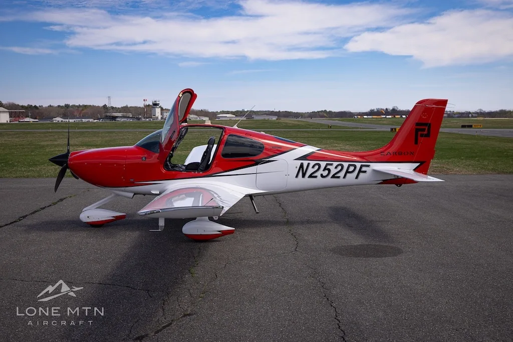 A side view of a red and white Cirrus SR20 aircraft with its doors open, parked on an airfield.