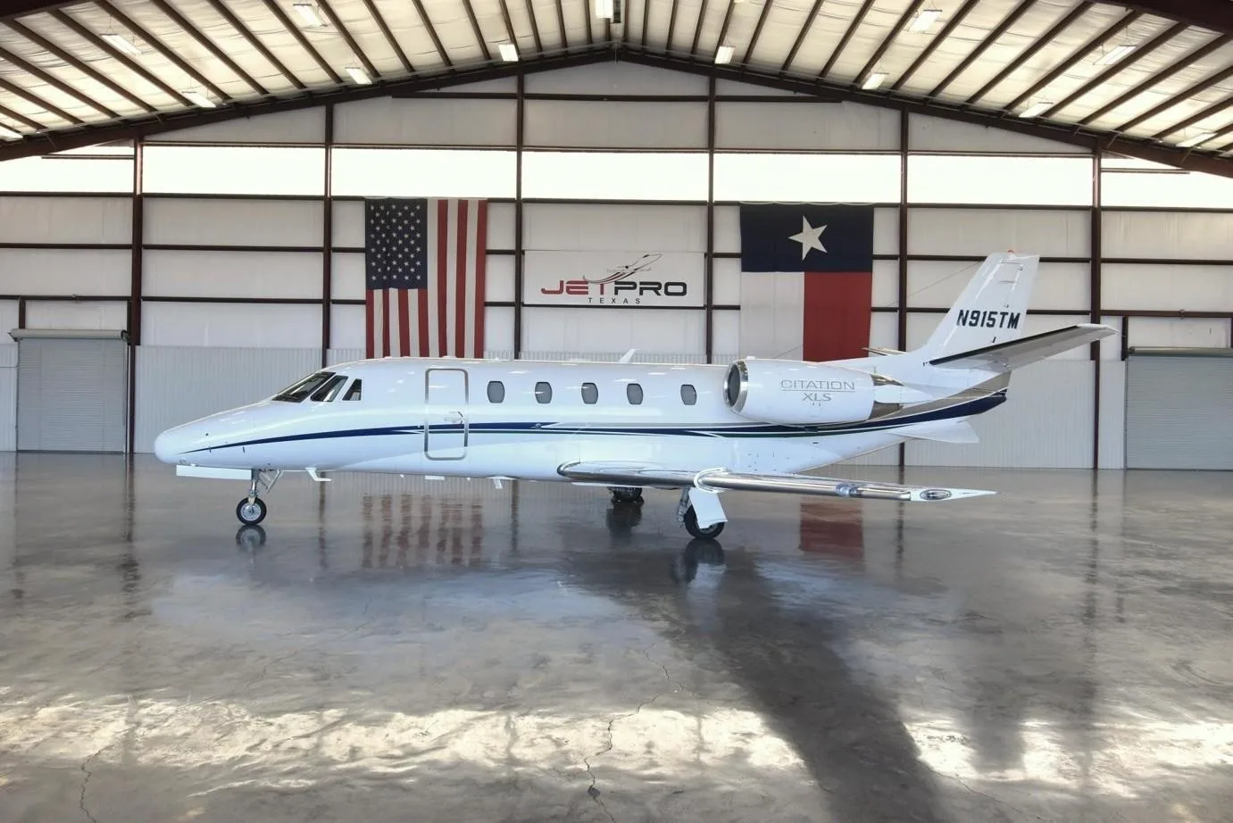 A white Cessna Citation XLS business jet with blue and silver stripes parked in a hangar with US and Texas flags.