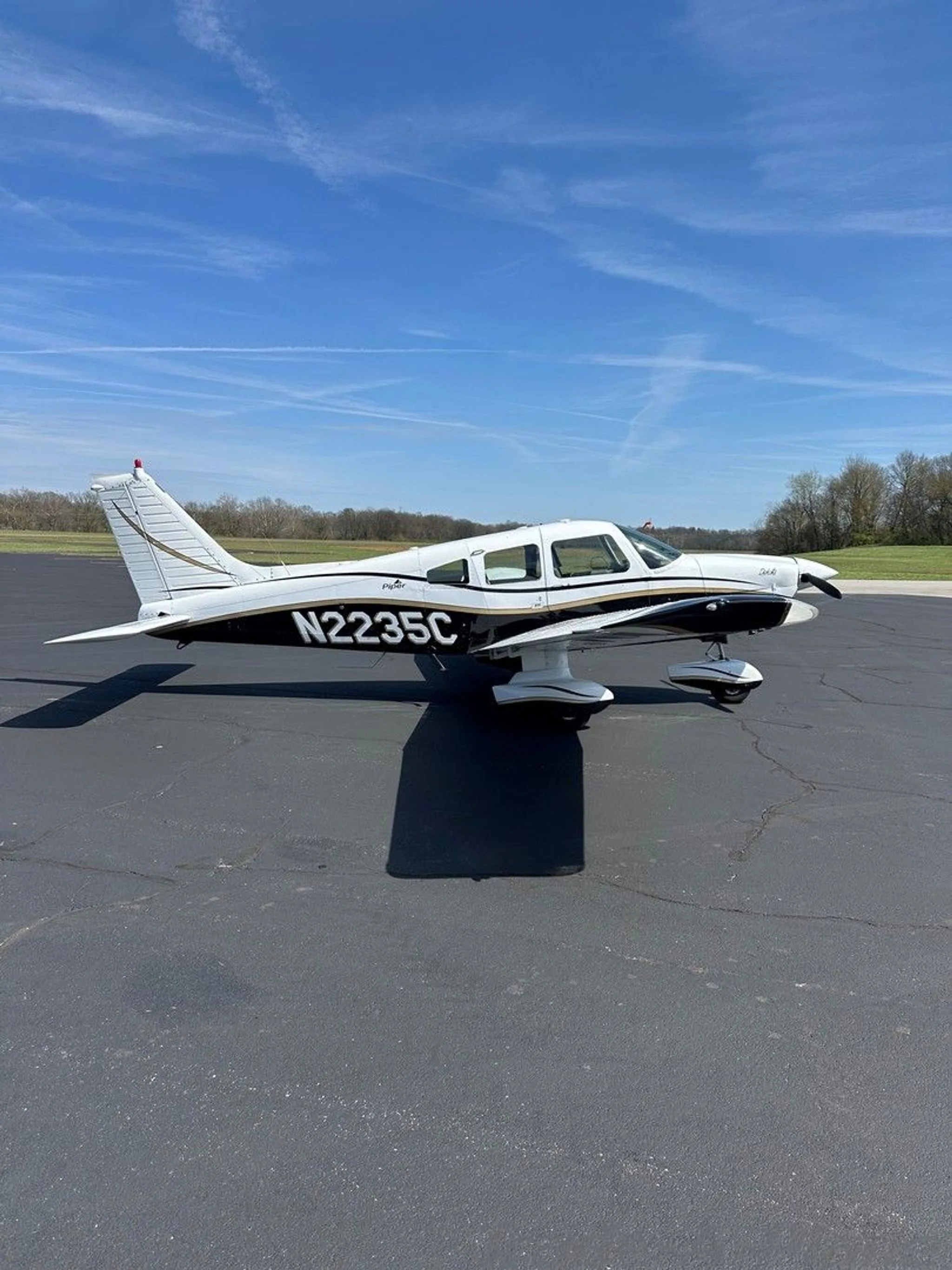 A white and black Piper aircraft with gold trim, registration N2235C, parked on an asphalt ramp under a clear blue sky.