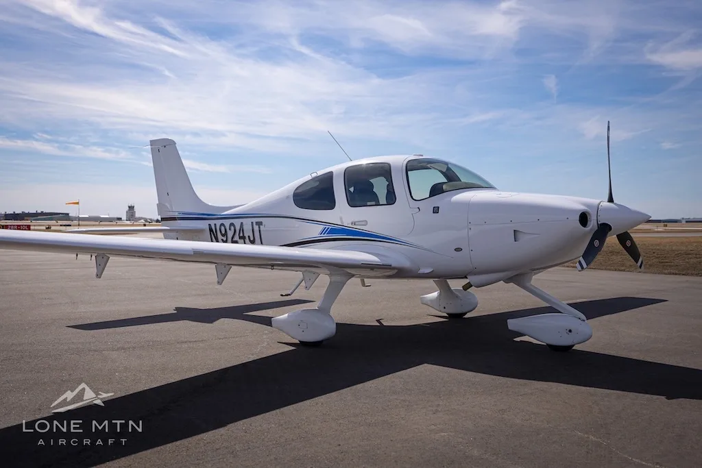 A white single-engine aircraft with blue and black stripes, tail number N924JT, parked on an airport tarmac under a partly cloudy sky.