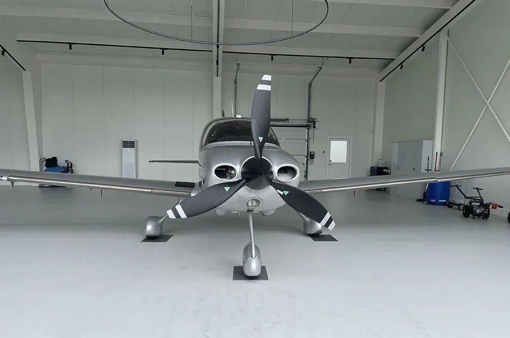 A head-on view of a silver Cirrus SR22 Turbo aircraft with a three-bladed propeller, parked inside a hangar.