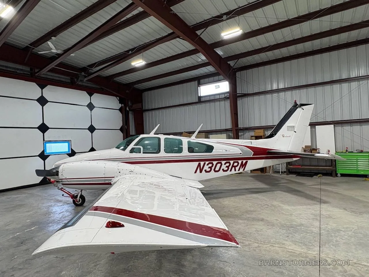 A white twin-engine propeller aircraft with red and grey stripes and tail number N303RM, viewed from the left side with the wing prominent, inside a hangar.