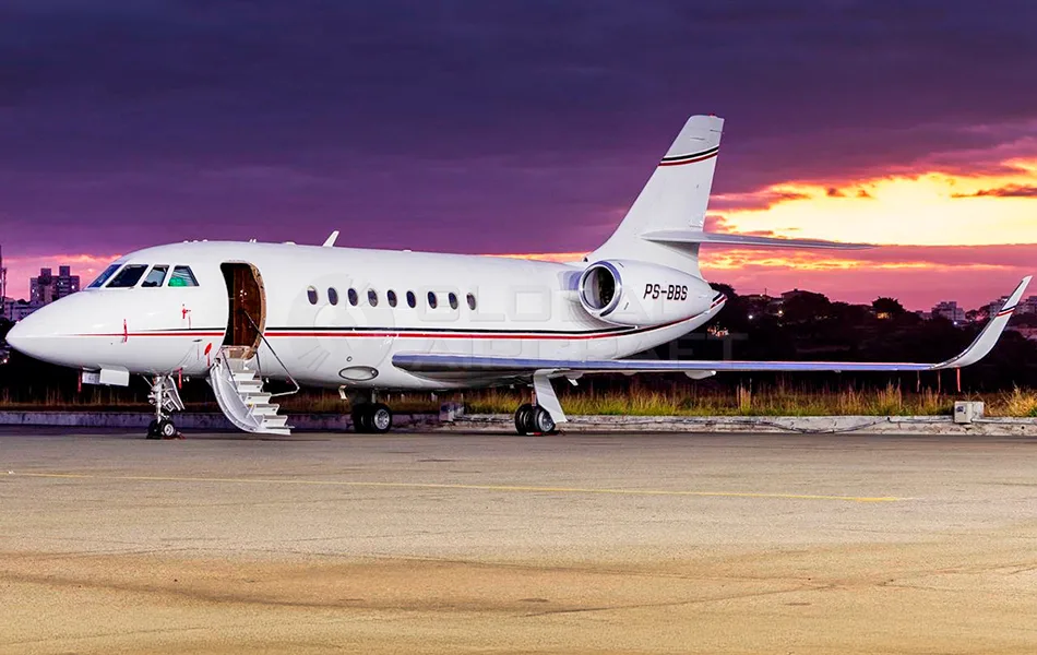 A white Dassault Falcon business jet with red and black stripes, registration PS-BBS, parked on a tarmac with its door open at sunset.