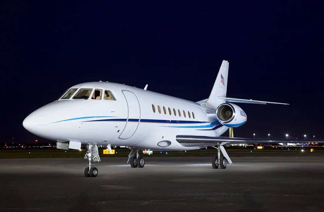 A white Dassault Falcon 900 business jet with blue stripes is parked on an airport tarmac at night.