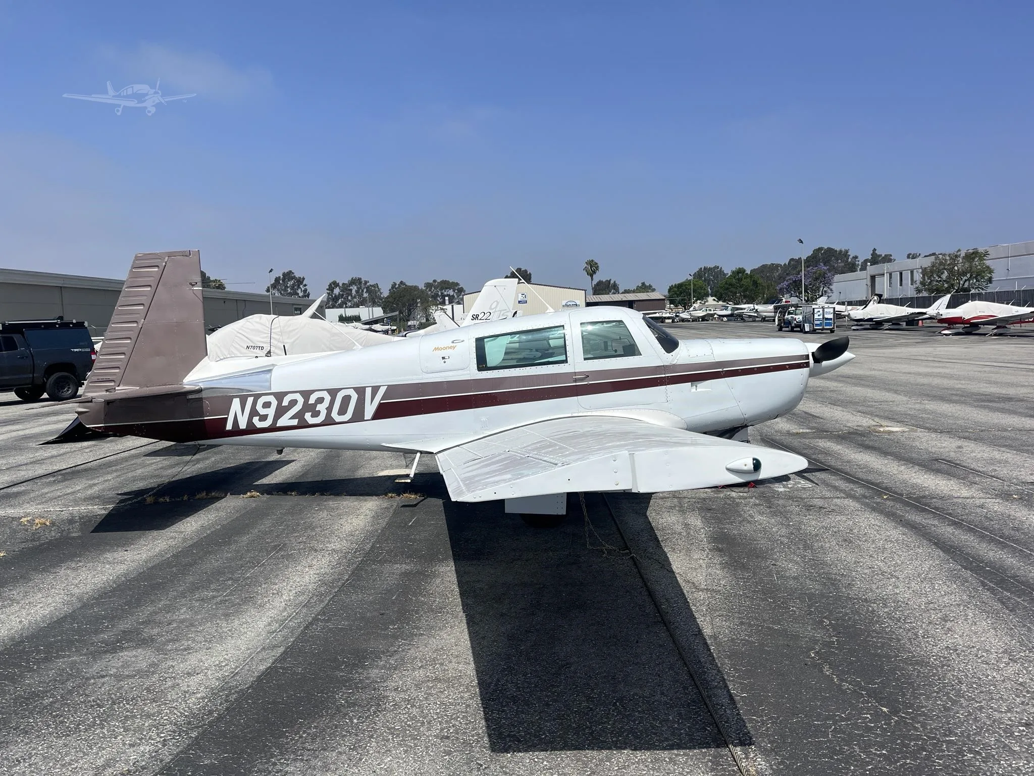 A white and brown Mooney M20 aircraft, registration N9230V, is parked on an asphalt tarmac at an airport on a sunny day.