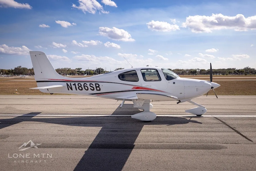 A side profile of the white N186SB single-engine aircraft with red and black stripes, parked on an airfield.