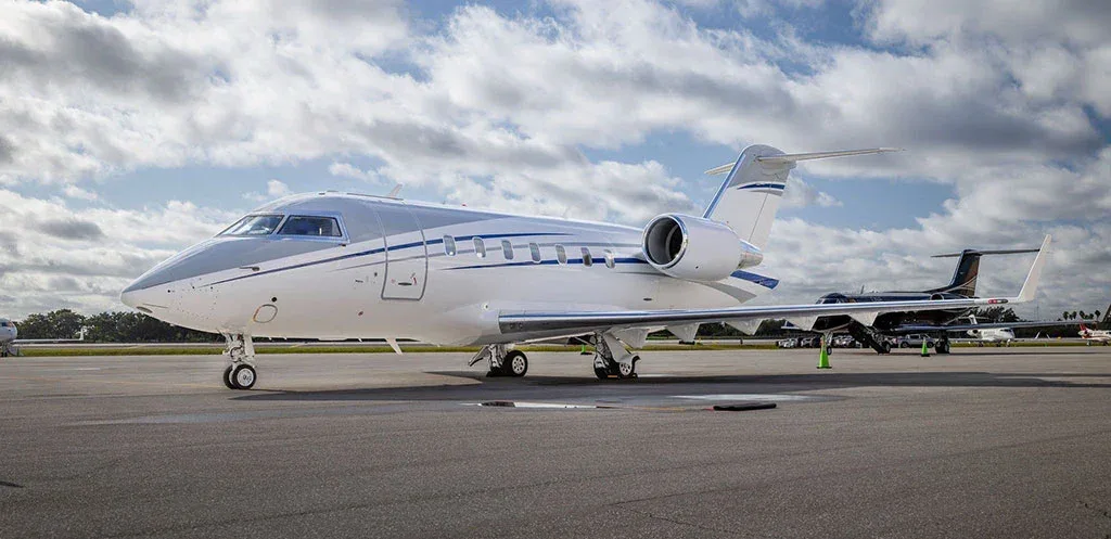 A white and grey Bombardier Challenger 605 business jet parked on an airport tarmac under a cloudy sky.