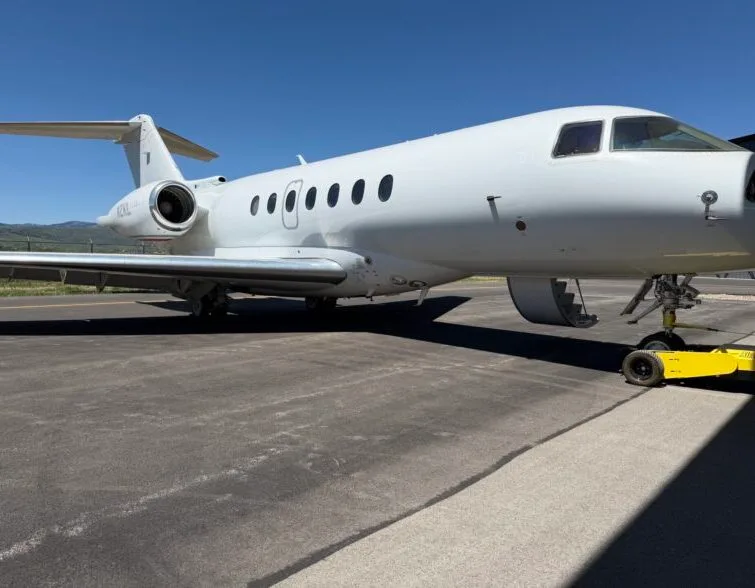 A white business jet, likely a Dassault Falcon, parked on an asphalt tarmac with mountains in the background under a clear blue sky.