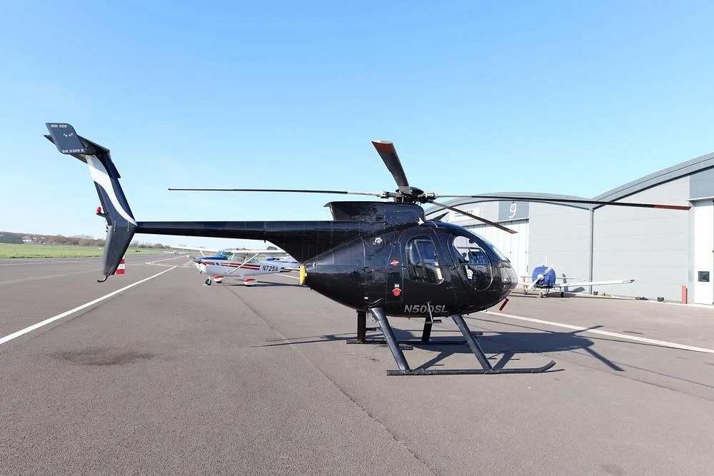 A black MD 500 helicopter (N500SL) is parked on an airfield tarmac in front of hangars.