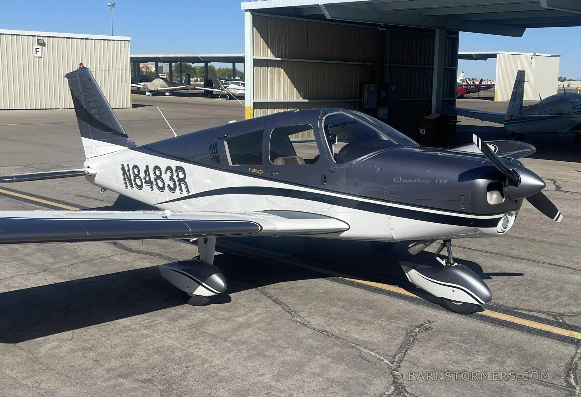 A Piper Cherokee 140 aircraft with a dark gray and white paint scheme, parked on an airfield next to a hangar.