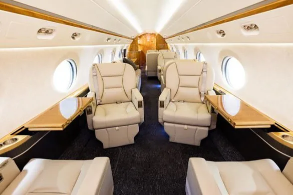 The luxurious interior of a private jet cabin with beige leather seats, wooden tables, and round windows.