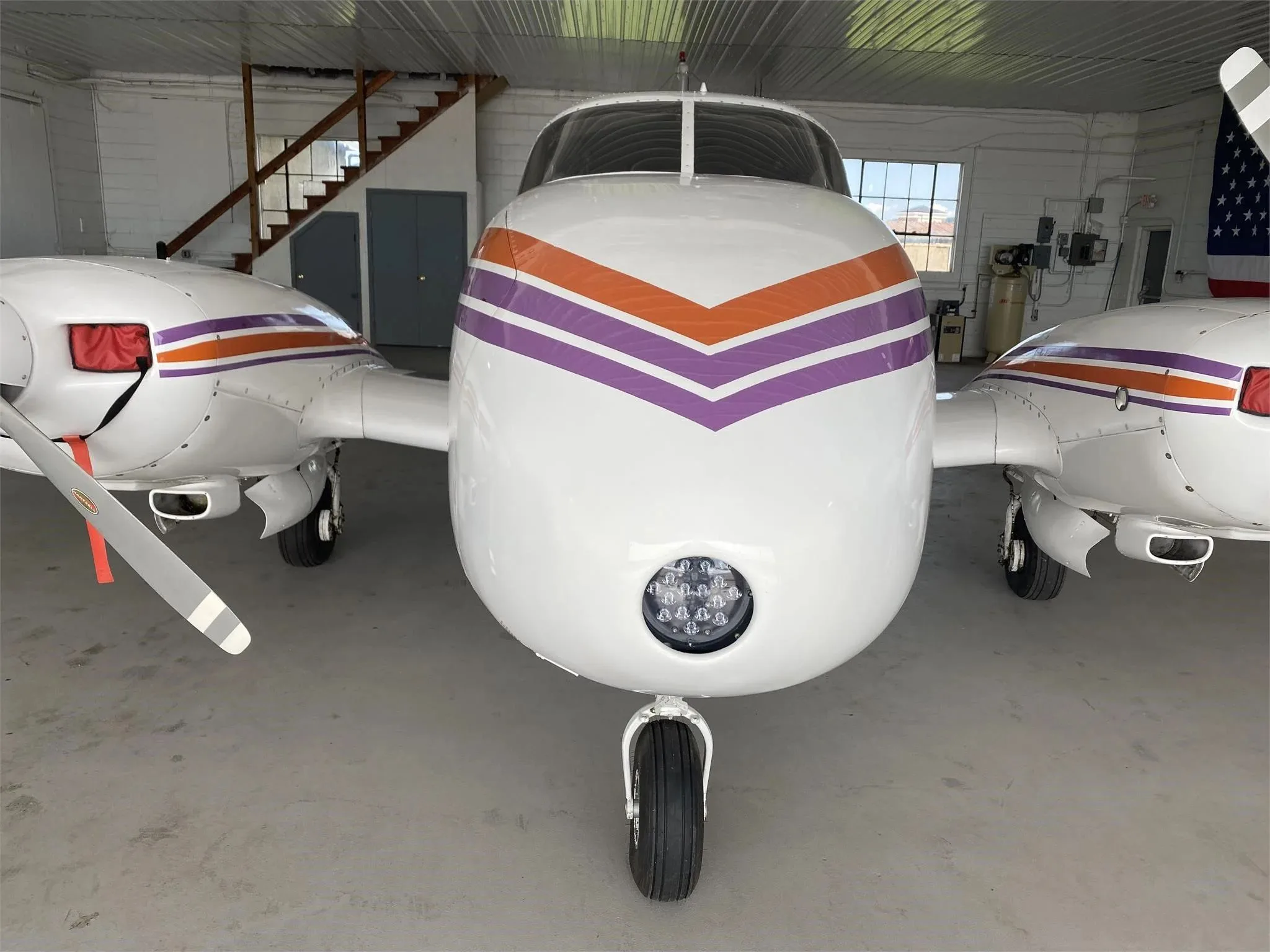A head-on view of the white Piper Aztec aircraft, highlighting its nose landing gear and orange and purple striped paint scheme.