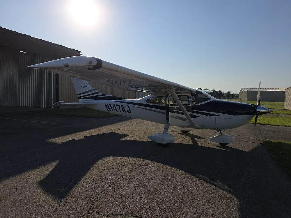 Side view of a white and blue Cessna 182 aircraft with registration N147AJ parked on an asphalt ramp under a sunny sky.