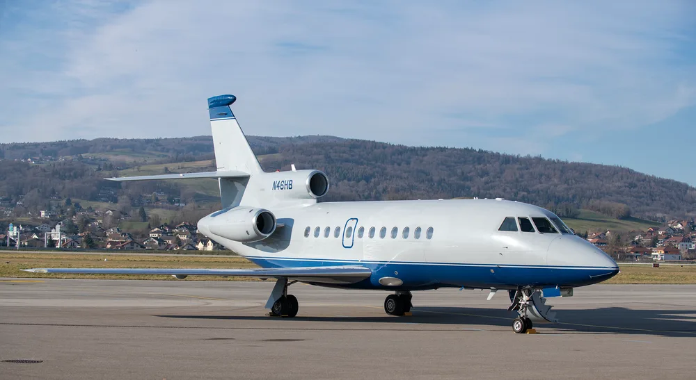 A Dassault Falcon 900EX business jet, registration N46HB, in white and blue livery, parked on an airport tarmac with hills in the background.