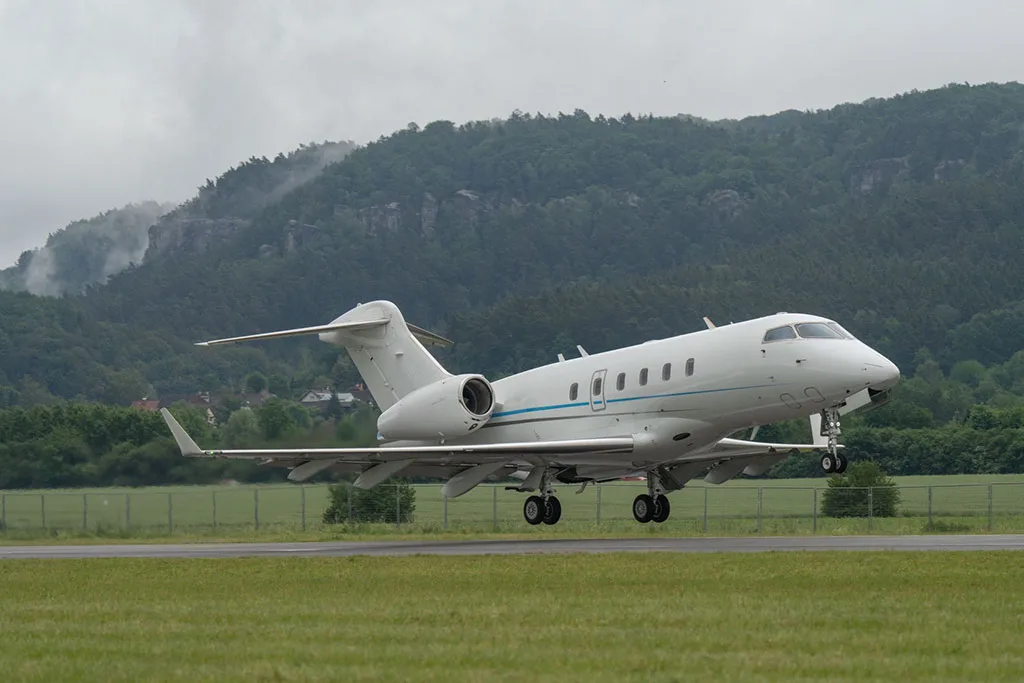 A white business jet with a blue stripe, seen from the side, with its landing gear extended, on a runway with green hills in the background.