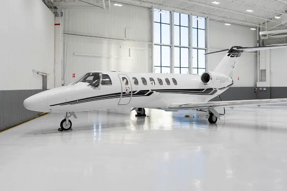 A white Cessna Citation CJ3+ business jet with black stripes is parked inside a brightly lit hangar.