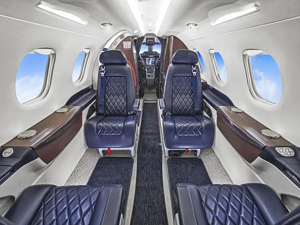 The aircraft cabin features two rows of dark blue quilted leather seats facing each other, with large windows and overhead lighting.