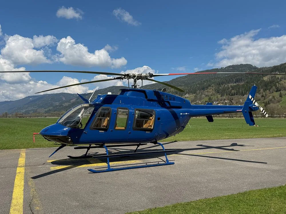 A blue Bell 505 helicopter sits on a tarmac with mountains and a blue sky in the background.