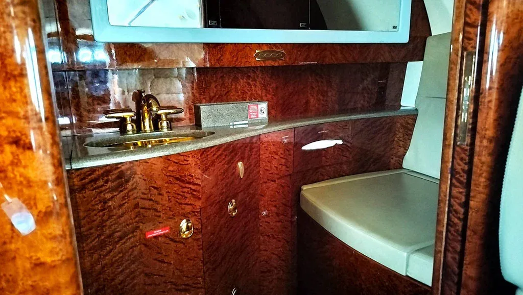 A private jet lavatory or galley area featuring a gold-colored sink faucet, a mirror, and rich burled wood paneling.
