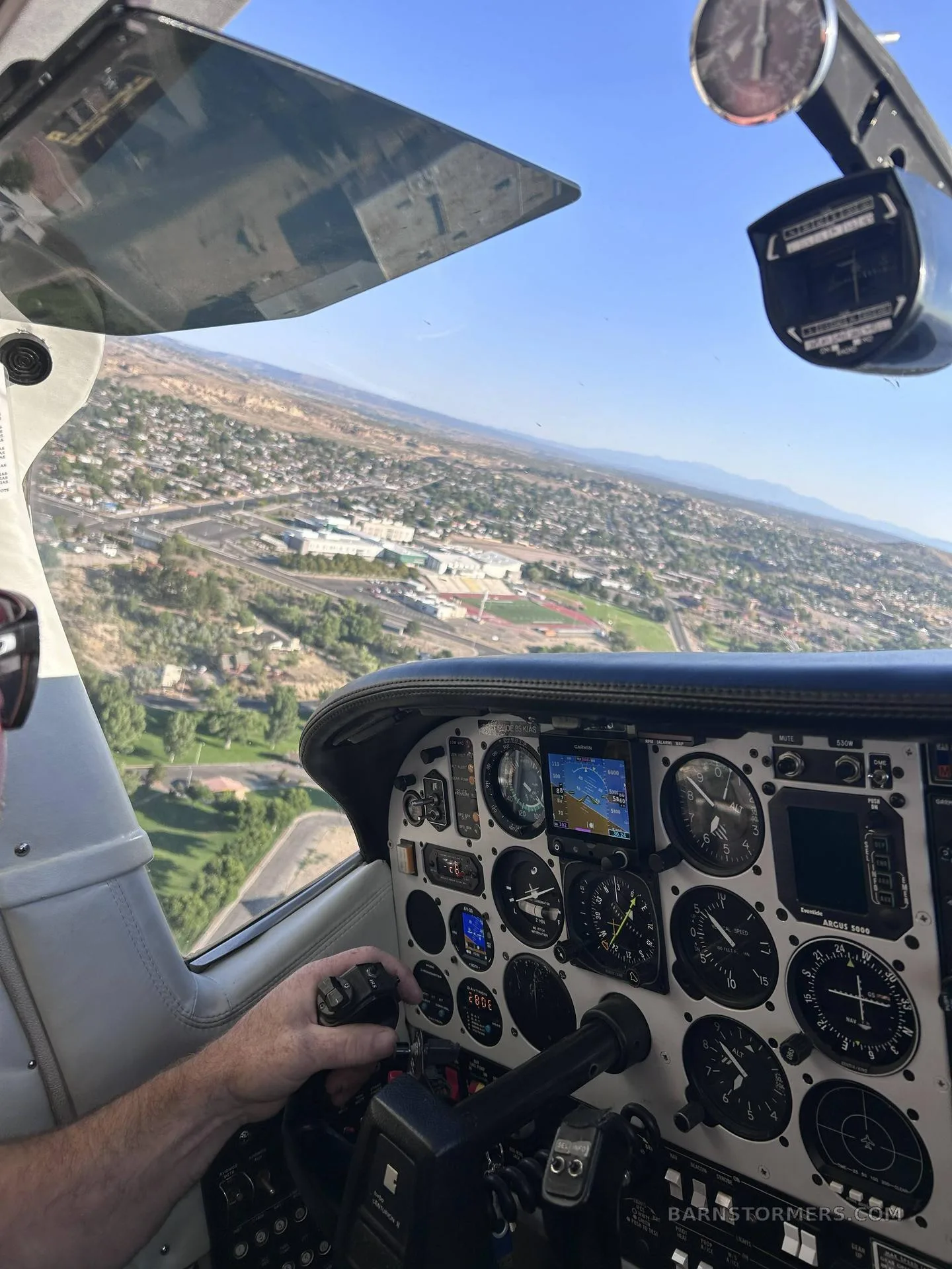 A pilot's view from the cockpit of a small aircraft, showing instruments and an urban landscape below.