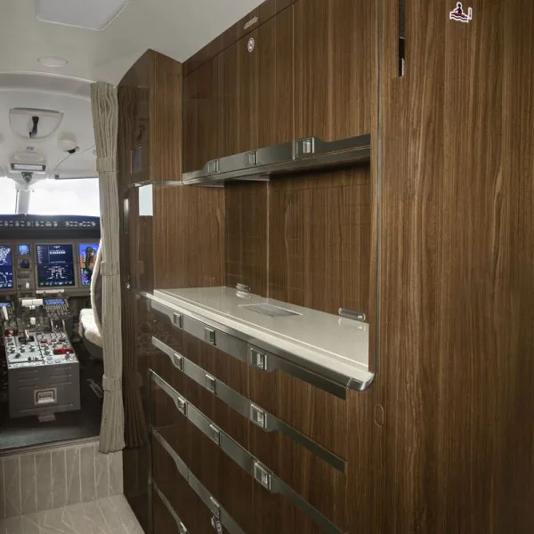 The galley area of a private jet with wood cabinetry and a counter, with the cockpit visible in the background.